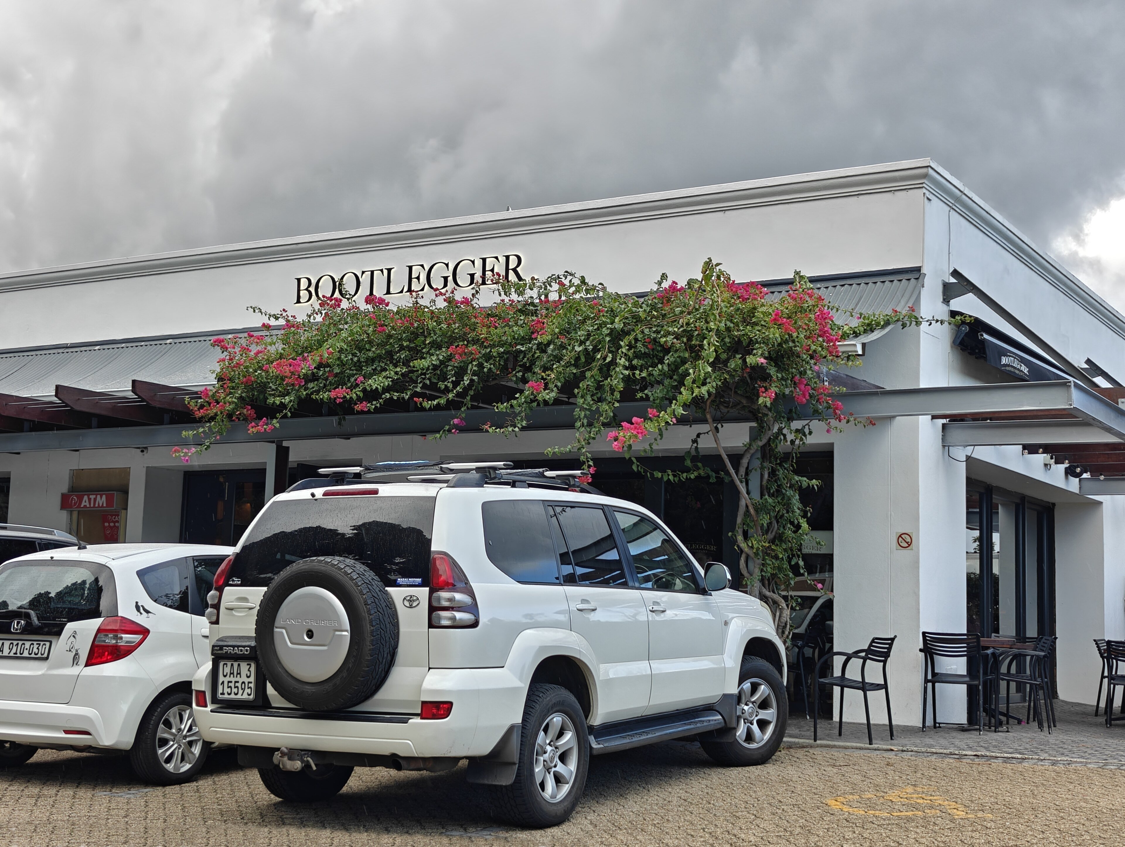 The front of a Bootlegger coffee shop in Tokai, Cape Town, with bougainvillea over the entrance and cars parked outside
