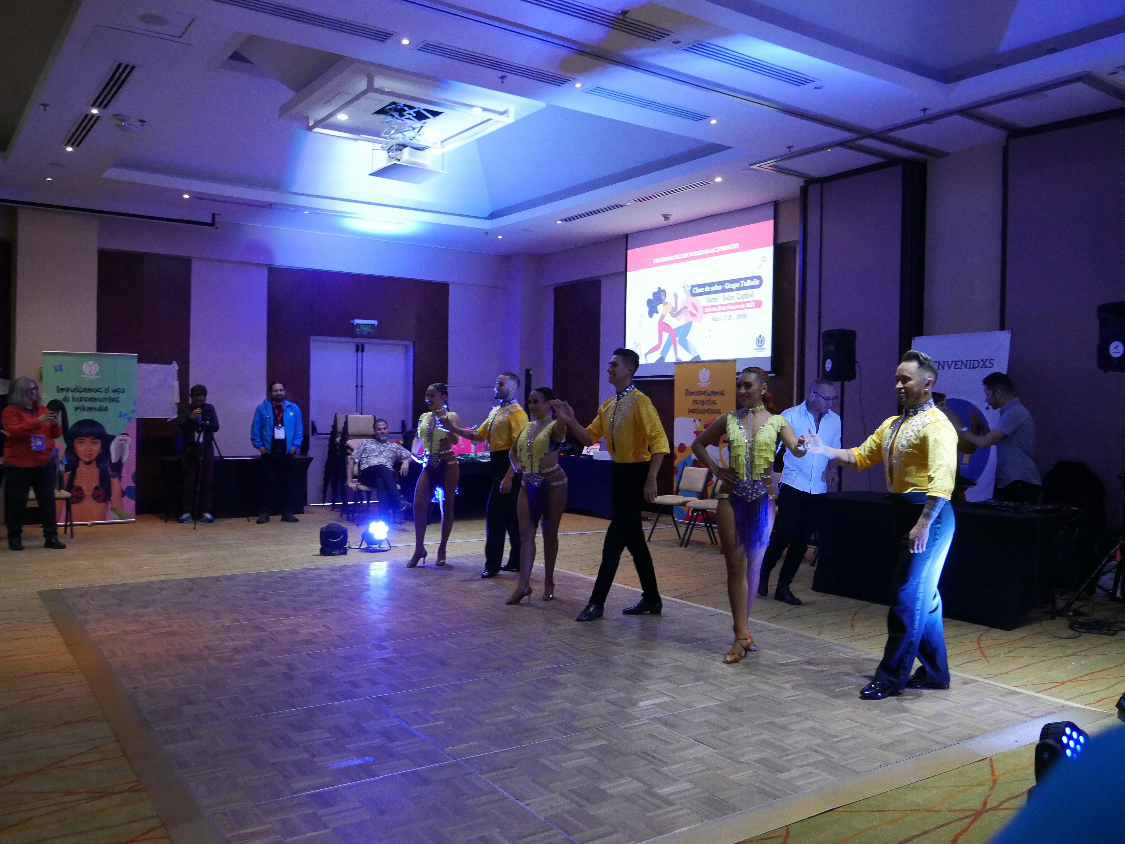 Salsa dancers in yellow and green costumes dancing in pairs across a ballroom floor under blue stage lights