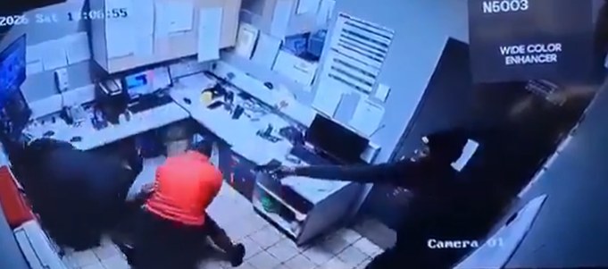 CCTV still from an office camera showing a man pointing a pistol, with a person in a red shirt crouching nearby