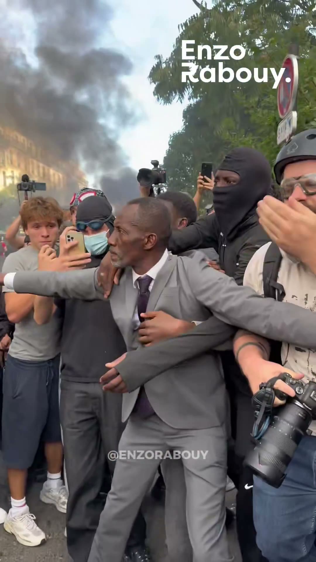 Bally Bagayoko in a grey suit stands with open arms in front of riot police and a crowd of young protesters in Paris