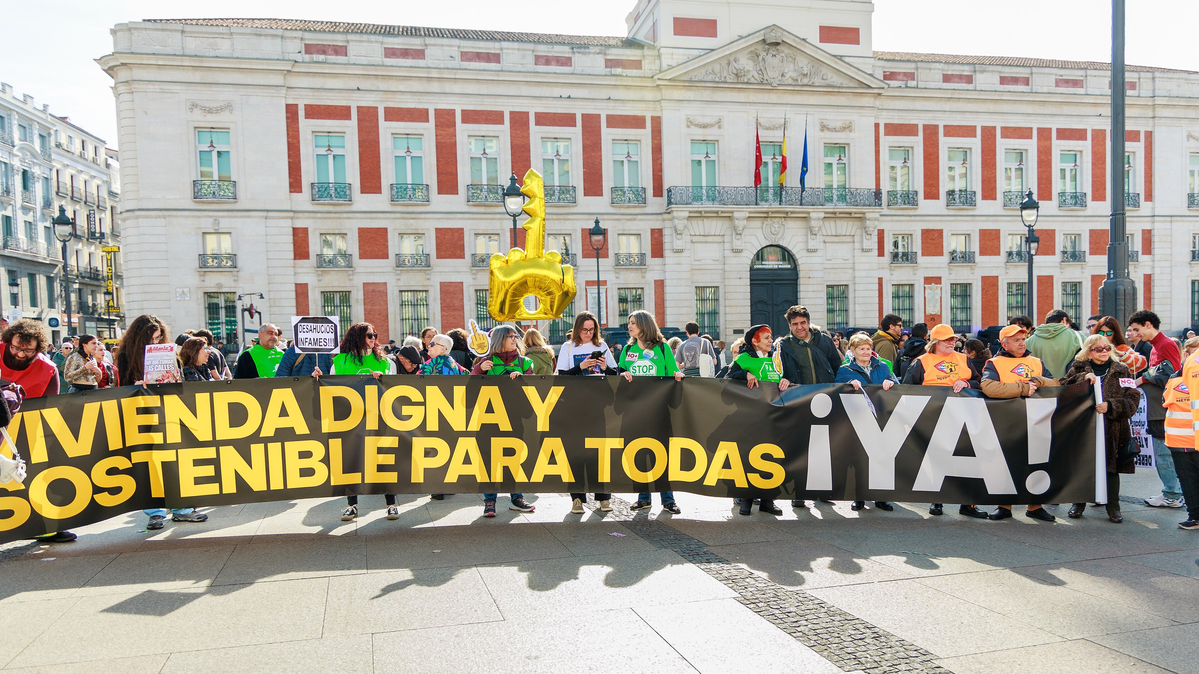 Housing protesters hold a long black and yellow banner in front of the red and white government building on Puerta del Sol in Madrid, with a giant gold key balloon above them