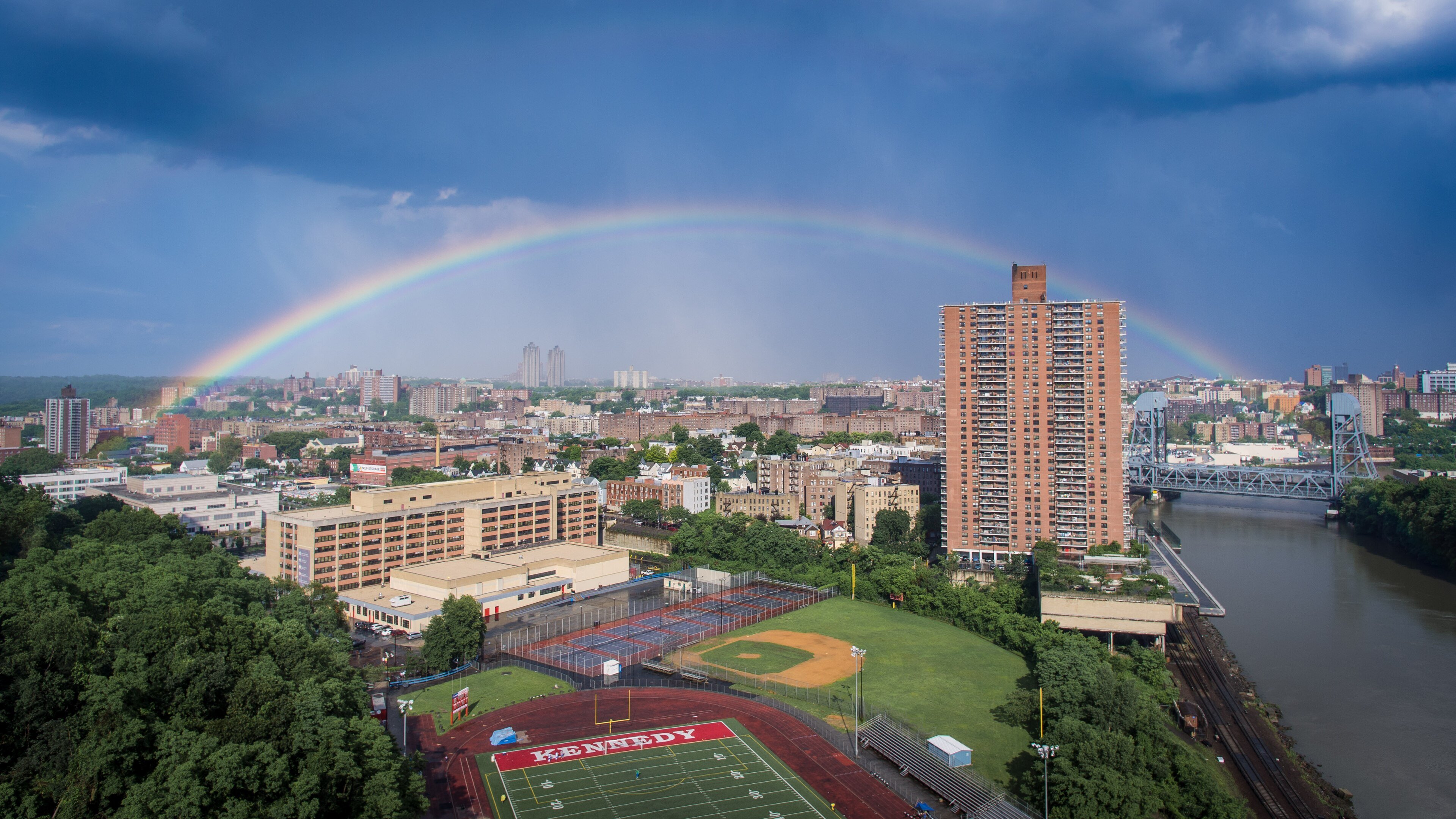 Aerial view of Marble Hill with a tall brick apartment tower, a high school sports field and the Harlem River under a rainbow