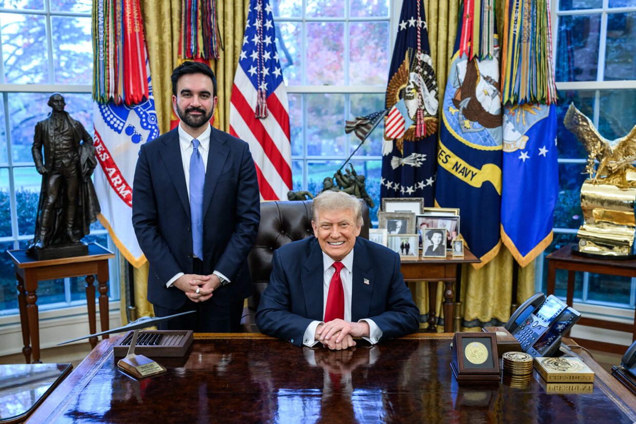 Zohran Mamdani standing beside President Donald Trump, who sits smiling at his desk in the Oval Office
