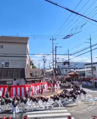 A huge wooden danjiri festival float coming down a Japanese street while teams of pullers in dark coats run across the junction