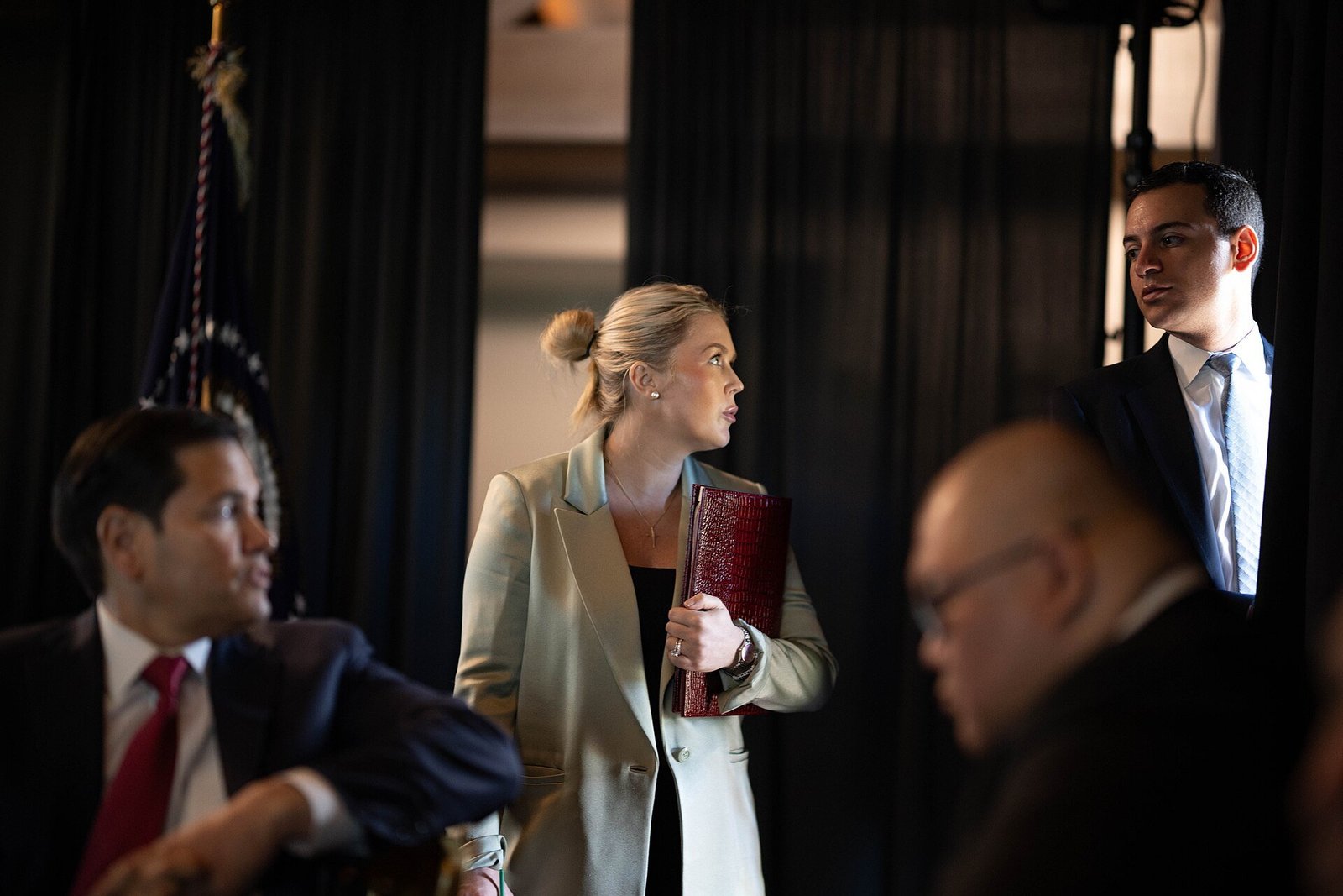 Karoline Leavitt holding a red folder at a briefing with Marco Rubio in the foreground