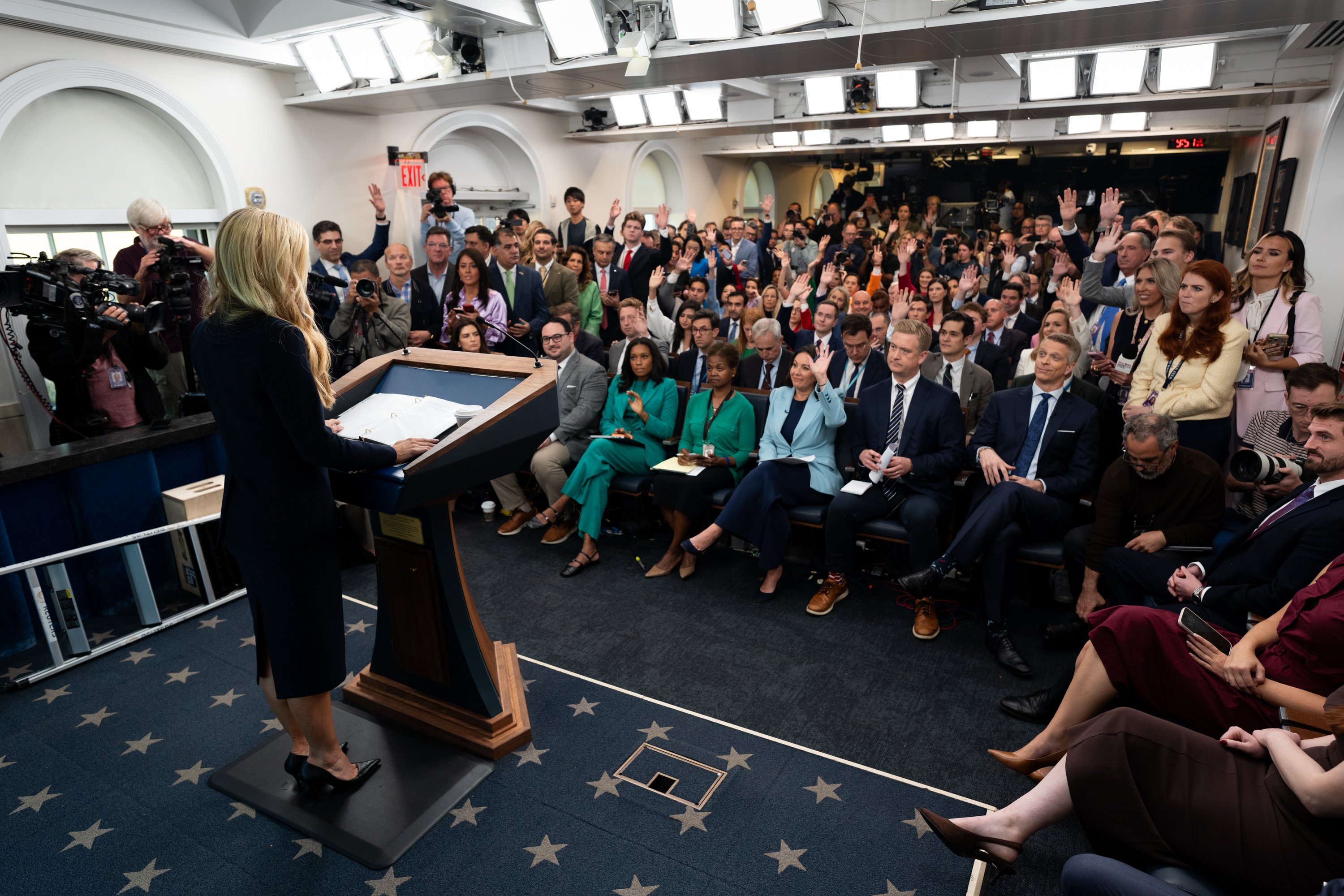 Katie Zacharia, seen from behind at the White House podium during her first press briefing, with reporters raising their hands