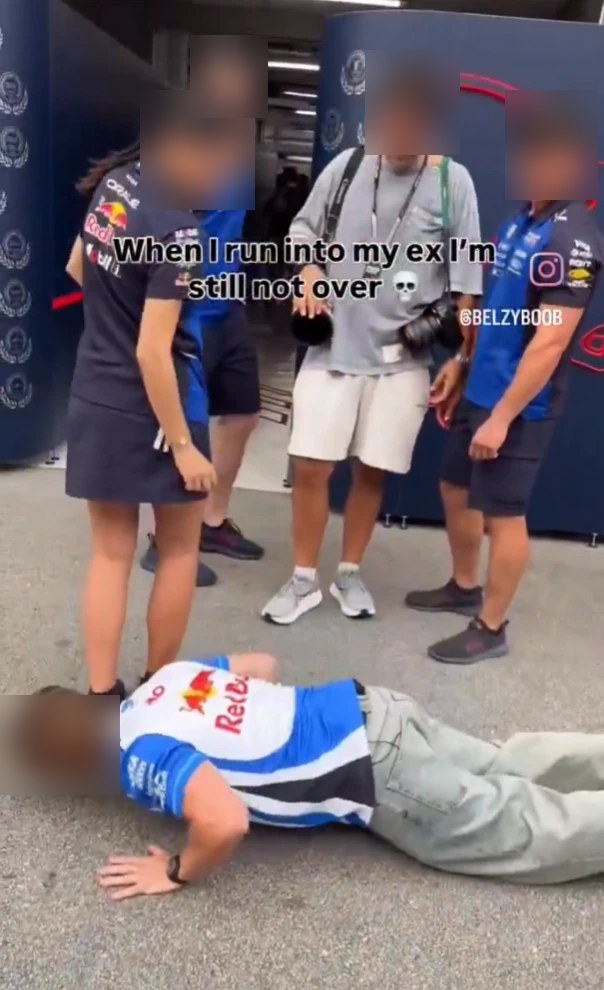 A man in a Red Bull shirt doing push-ups on the paddock floor while a female staff member and three men look on
