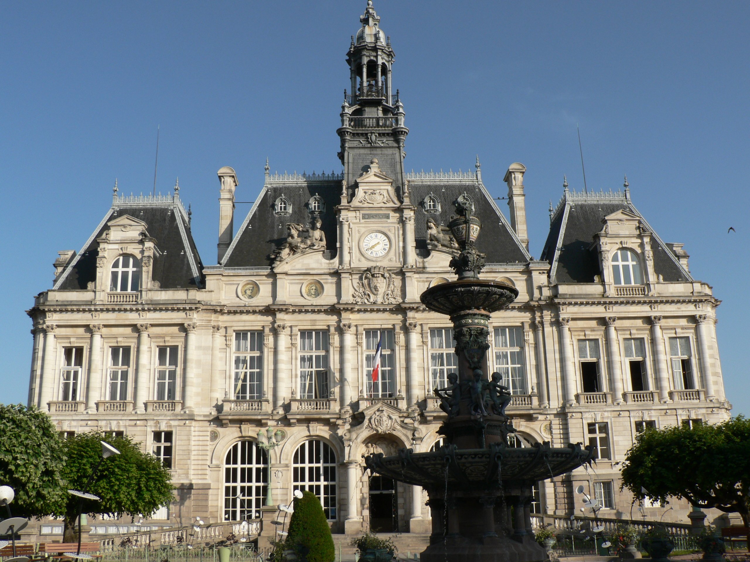 Limoges town hall in France with its stone front, slate roofs, clock and tall bell tower, behind a bronze fountain, before the fire