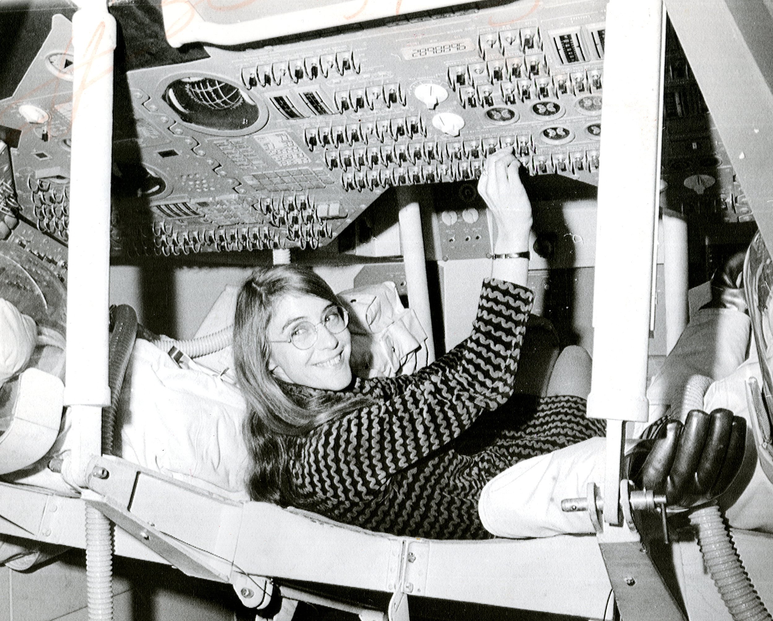 Margaret Hamilton sitting at the controls of an Apollo command module simulator, a panel of switches in front of her