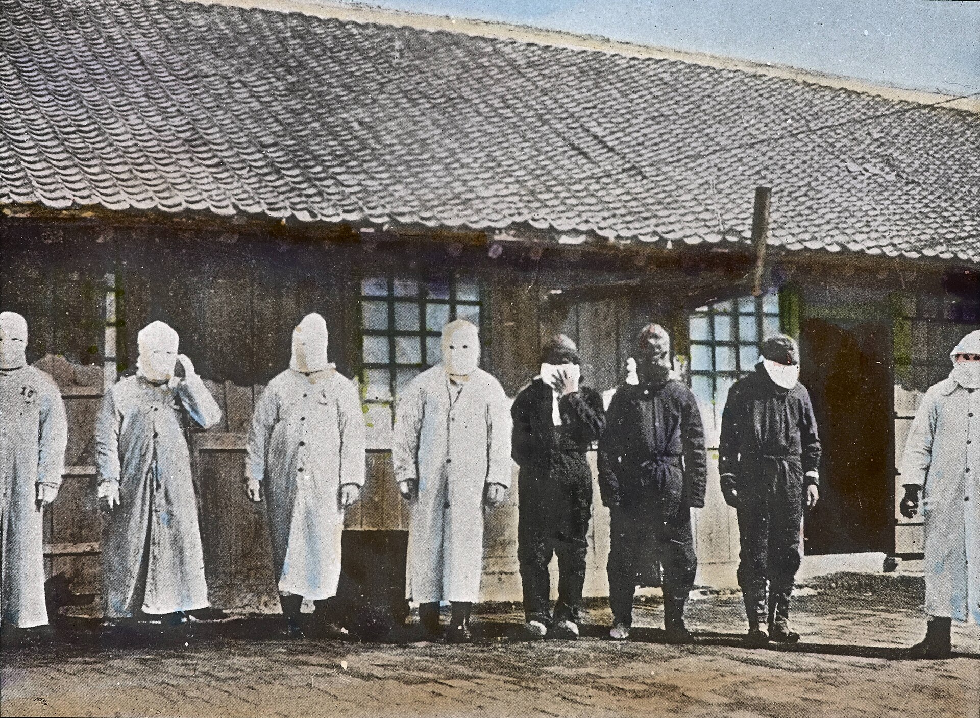 Plague workers in long white coats and cloth face masks standing outside a wooden hospital in Mukden, Manchuria, in 1911