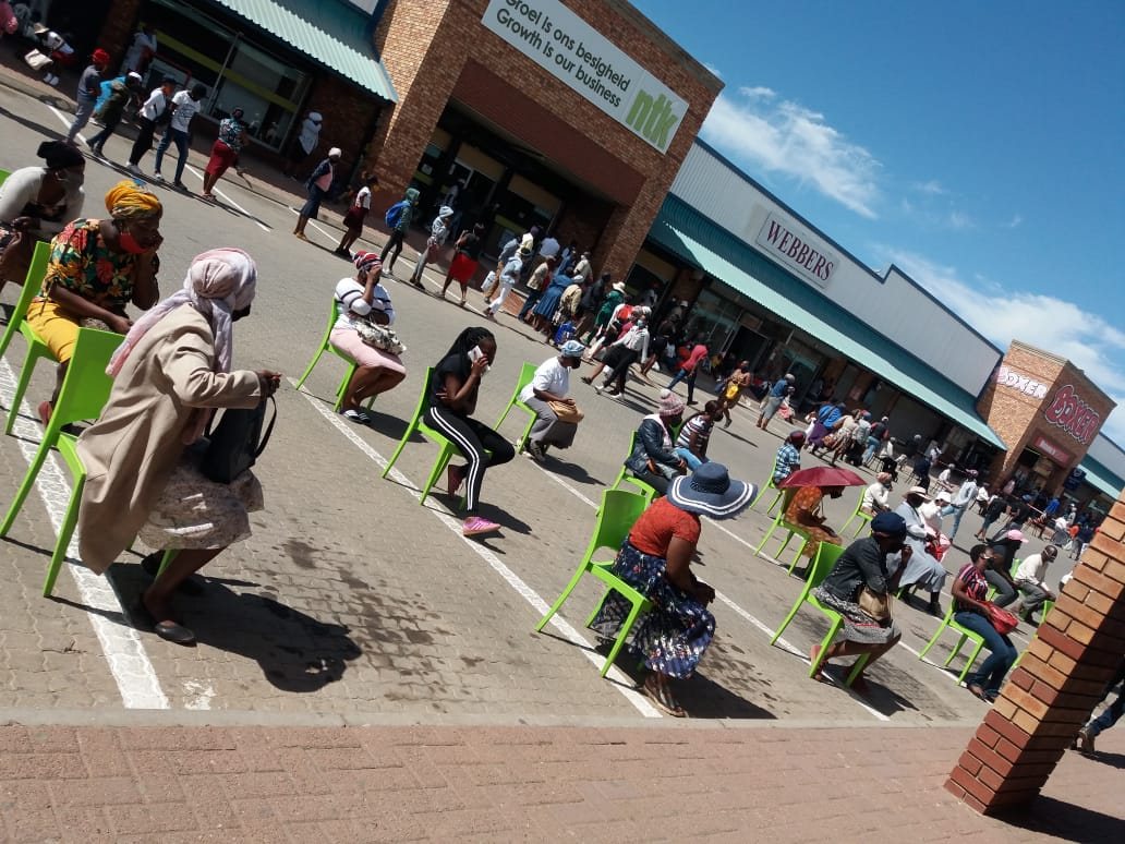 People waiting in a long line outside a social grant office, with plastic chairs set out for the elderly