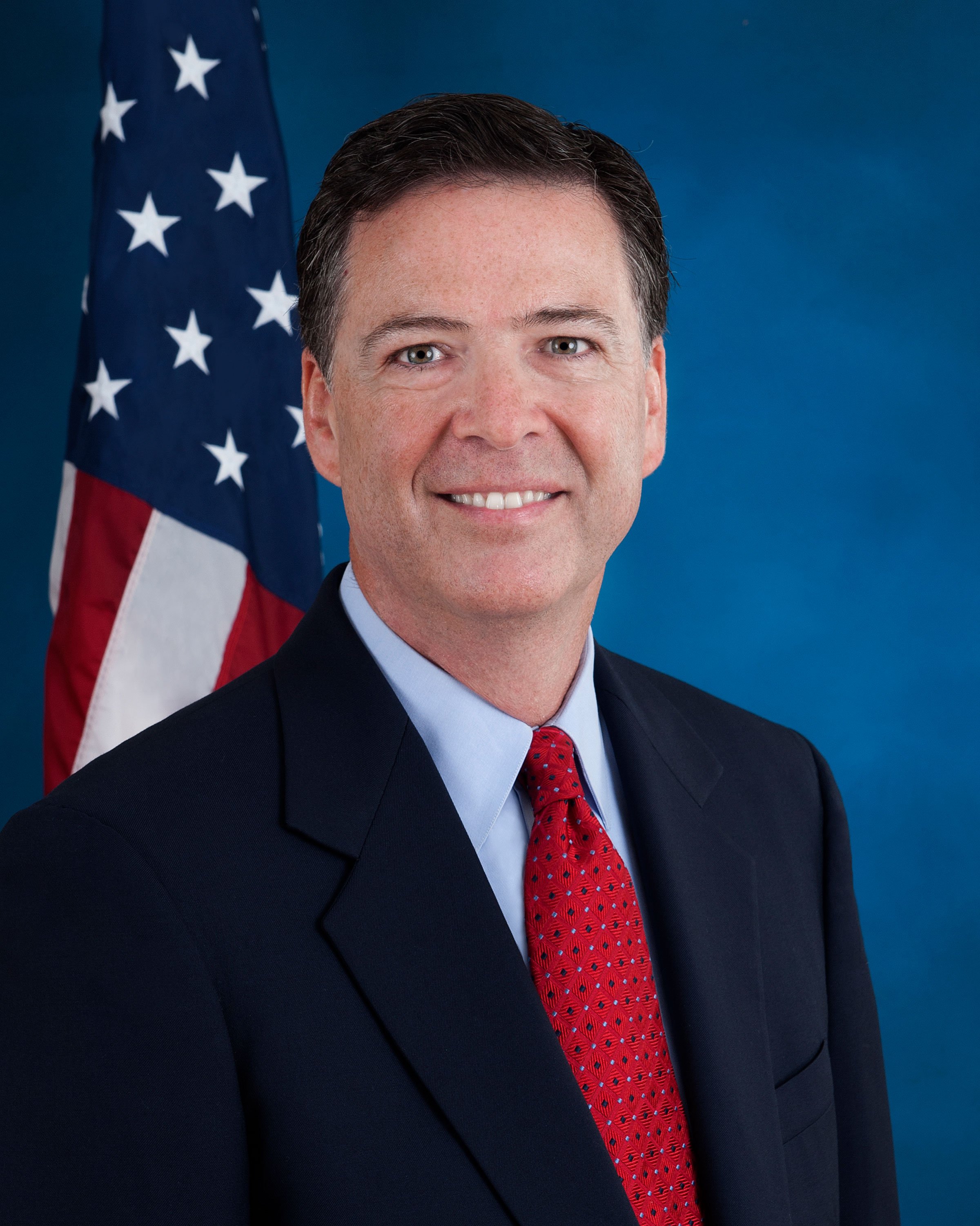 James Comey in a dark suit and red tie in front of an American flag
